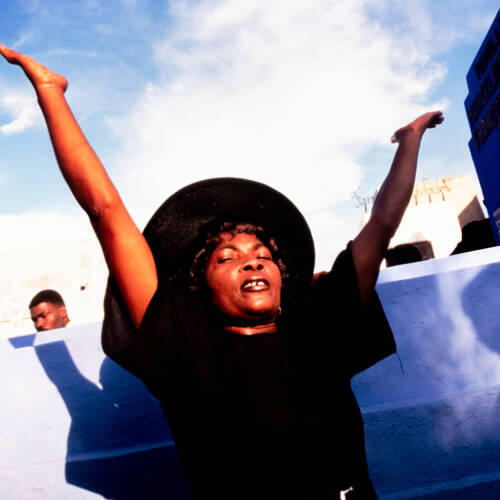 Color photograph of a woman wearing all black raising her hands towards the sky.